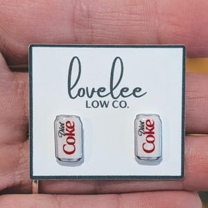 May include: A pair of stud earrings shaped like miniature silver and white Diet Coke cans with red lettering. The earrings are attached to a white rectangular display card featuring the black text lovelee LOW CO. centered above the earrings.