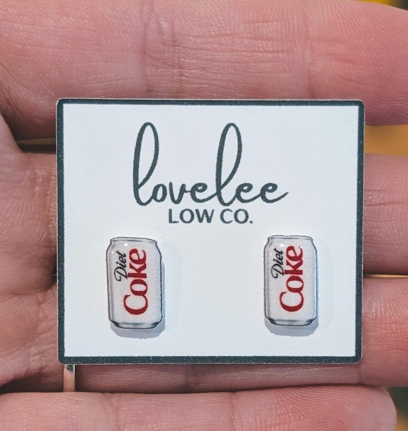 May include: A pair of stud earrings shaped like miniature silver and white Diet Coke cans with red lettering. The earrings are attached to a white rectangular display card featuring the black text lovelee LOW CO. centered above the earrings.