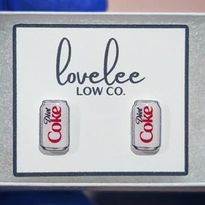 May include: A pair of stud earrings shaped like silver Diet Coke cans with red and black text, displayed on a white card featuring the black printed text lovelee LOW CO. The earrings are positioned side by side on the rectangular display card.
