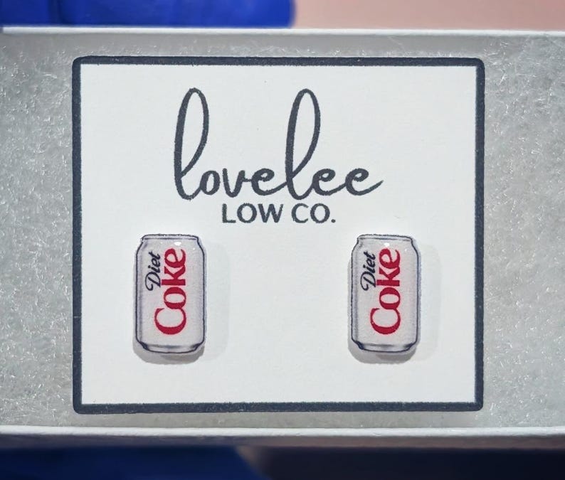May include: A pair of stud earrings shaped like silver Diet Coke cans with red and black text, displayed on a white card featuring the black printed text lovelee LOW CO. The earrings are positioned side by side on the rectangular display card.