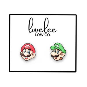 Puede incluir: Pendientes de botón con las caras de Mario y Luigi de Super Mario Bros. Mario tiene un sombrero rojo y Luigi uno verde. Los pendientes están en una tarjeta blanca con el texto "lovelee LOW CO."