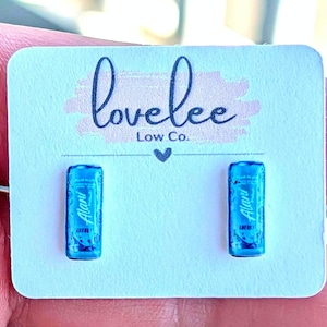 May include: A pair of stud earrings shaped like miniature blue energy drink cans, each featuring the Alani Nu logo. The earrings are mounted on a white rectangular display card printed with the text lovelee Low Co. and a small heart icon.