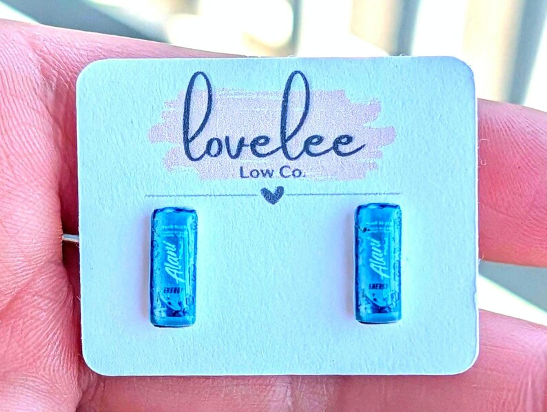 May include: A pair of stud earrings shaped like miniature blue energy drink cans, each featuring the Alani Nu logo. The earrings are mounted on a white rectangular display card printed with the text lovelee Low Co. and a small heart icon.