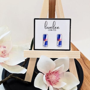 May include: A pair of stud earrings featuring miniature Red Bull energy drink cans on a white display card with the text lovelee LOW CO. printed in black. The earrings are mounted on a small wooden easel stand.