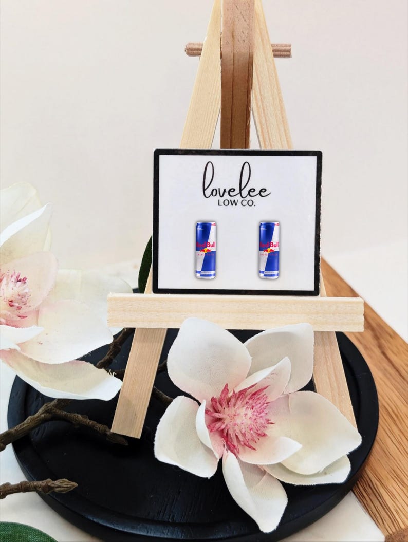 May include: A pair of stud earrings featuring miniature Red Bull energy drink cans on a white display card with the text lovelee LOW CO. printed in black. The earrings are mounted on a small wooden easel stand.