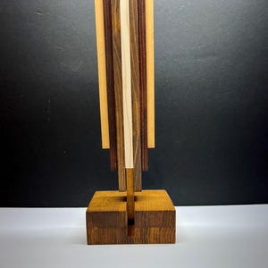 Może przedstawiać: Drewniana rzeźba z prostokątną podstawą i pionowym wzorem. Rzeźba charakteryzuje się różnorodnością odcieni drewna, w tym jasnymi, średnimi i ciemnymi brązami oraz centralnym paskiem z naturalnego drewna. Projekt jest geometryczny i abstrakcyjny.