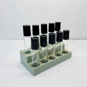 May include: A collection of clear glass roller bottles with black caps, arranged in a tiered, light green rectangular holder. The bottles are empty and ready for essential oils or perfumes. The holder is made of a solid material.