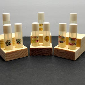 May include: A collection of lip balm tubes with white caps, displayed in wooden holders. The tubes are clear with decorative patterned labels. The holders are made of natural wood and have a stepped design.