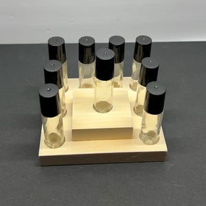 May include: A wooden display stand holding ten clear glass roller bottles with black caps. The bottles are arranged in a square pattern around a central bottle. The stand is made of light-colored wood.