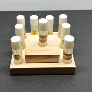 May include: A wooden peg game with a square base and a smaller square in the center. Ten clear tubes with white caps are arranged around the squares. The tubes have decorative patterns and contain a liquid.