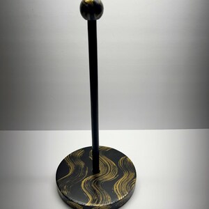 May include: A black and gold decorative stand with a round base and a tall, slender pole. The base features a black background with gold wave-like patterns. The top of the pole has a small, round, marbled gold and black accent.