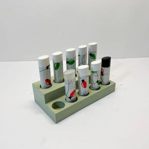 May include: A light green wooden display stand holding ten cylindrical lip balm tubes. The tubes are white with a festive design of red and green Christmas lights. One tube has a black cap.