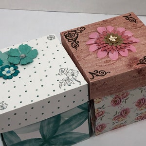May include: Two decorative boxes with floral designs. The top box is brown with a pink flower and black floral accents. The bottom box is white with green polka dots and green flowers. Both boxes have a floral pattern on the bottom.