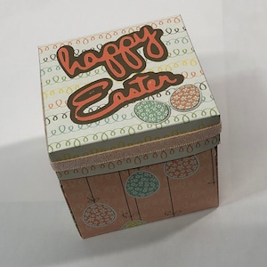 May include: A square gift box with a pink, white, and green pattern. The top of the box says "happy Easter" in brown lettering. The box is decorated with a pink ribbon.