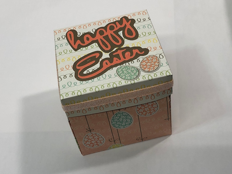 May include: A square gift box with a pink, white, and green pattern. The top of the box says "happy Easter" in brown lettering. The box is decorated with a pink ribbon.