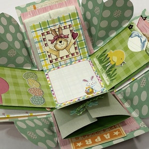 May include: A green and pink explosion box with a bunny, chick, and Easter egg designs. The box has a green and white checkered pattern and a polka dot pattern. The box is open and the inside is visible. The box has a message that says "Apr. 12".
