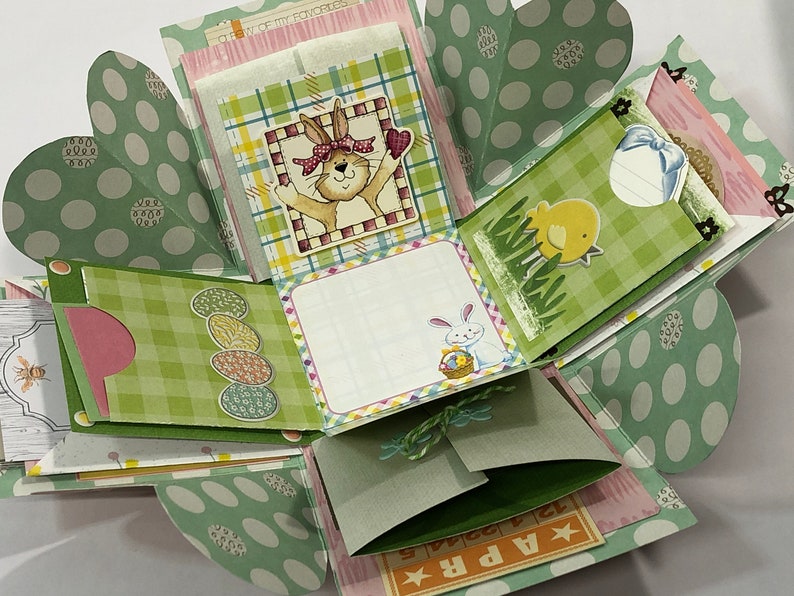 May include: A green and pink explosion box with a bunny, chick, and Easter egg designs. The box has a green and white checkered pattern and a polka dot pattern. The box is open and the inside is visible. The box has a message that says "Apr. 12".