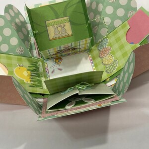 May include: A green and pink explosion box with a checkered pattern and a bunny theme. The box has a variety of cutouts and flaps, and is decorated with images of bunnies, chicks, and eggs. The box is tied with a green ribbon.