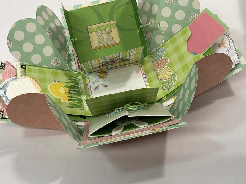 May include: A green and pink explosion box with a checkered pattern and a bunny theme. The box has a variety of cutouts and flaps, and is decorated with images of bunnies, chicks, and eggs. The box is tied with a green ribbon.