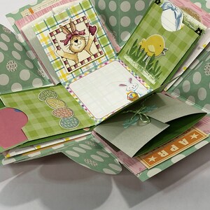May include: A green and white polka dot explosion box with a bunny, chick, and egg designs. The box has a green and white checkered pattern and a pink and white patterned paper inside. The box is decorated with a green ribbon and a tag that says "APR".