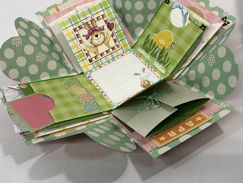 May include: A green and white polka dot explosion box with a bunny, chick, and egg designs. The box has a green and white checkered pattern and a pink and white patterned paper inside. The box is decorated with a green ribbon and a tag that says "APR".