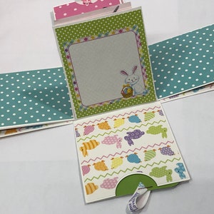 May include: A green, white, and pink Easter-themed paper craft box with a bunny and eggs design. The box is folded open to reveal a white interior with a blank space for writing. The box has a green pull tab.