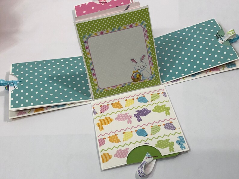 May include: A green, white, and pink Easter-themed paper craft box with a bunny and eggs design. The box is folded open to reveal a white interior with a blank space for writing. The box has a green pull tab.