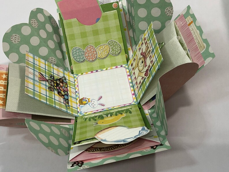 May include: A green and white explosion box with a bunny, eggs, and a chick design. The box has a checkered pattern and polka dots. The box is open and the inside is visible. The box is decorated with a variety of Easter-themed images.