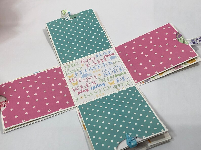 May include: A pink and blue patterned paper box with a white background. The box is decorated with the words "Easter", "Spring", "Flowers", "Rain", "Happy", "Show", "Play", "Wers", "Shower", and "Easter".