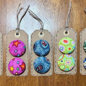 May include: Six sets of fabric covered buttons on brown card stock tags with twine. The buttons are in various patterns including polka dots, stars, paisley, and floral designs.