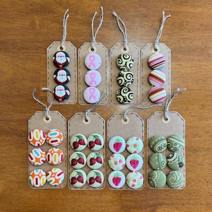 May include: Nine sets of three fabric buttons on brown card stock tags. The buttons feature various designs including Santa Claus, pink ribbons, swirls, stripes, strawberries, and paisley patterns.