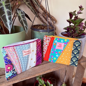 May include: Two patchwork zippered pouches in various fabrics, including floral and geometric patterns. One pouch features the text "bisou magique", the other "HO, FUCK" on a pink label. They are displayed on a wooden shelf, surrounded by plants.