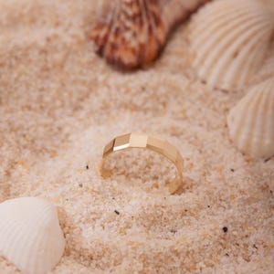 May include: A gold-colored ring with a geometric design, resting on a bed of sand. The ring's surface is segmented into rectangular facets. Seashells are scattered around the ring. The ring is a jewelry item.