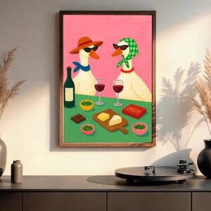 May include: A framed art print depicts two geese wearing sunglasses and accessories, enjoying wine and snacks. The artwork features a pink background, a green table, and a wooden frame. The word "SASSY" is on a red box.