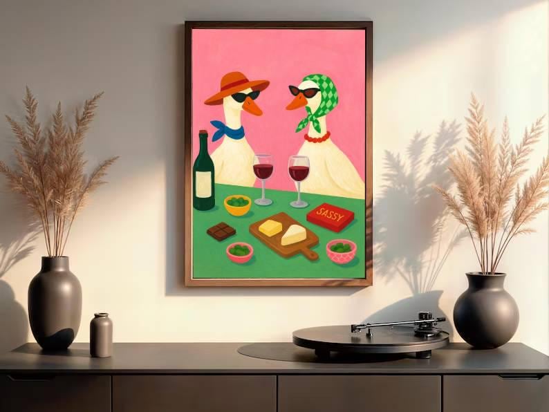 May include: A framed art print depicts two geese wearing sunglasses and accessories, enjoying wine and snacks. The artwork features a pink background, a green table, and a wooden frame. The word "SASSY" is on a red box.