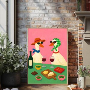 May include: A framed art print featuring two geese wearing sunglasses and accessories, enjoying wine and snacks. The geese are set against a pink background, with a green table displaying wine glasses, cheese, and olives. A red box reads "SASSY".