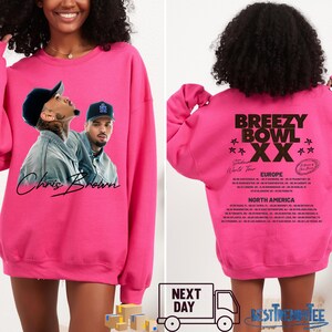 May include: A bright pink sweatshirt featuring a graphic of Chris Brown on the front. The back of the sweatshirt displays the text "Breezy Bowl XX" with a list of tour dates for Europe and North America.