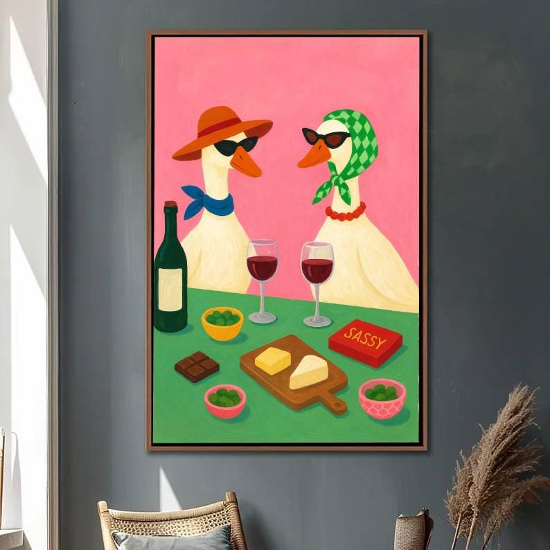 May include: A framed artwork depicts two geese wearing sunglasses and accessories, enjoying wine and snacks. The geese are set against a pink background, with a green table featuring wine glasses, cheese, olives, and a box labeled "SASSY".