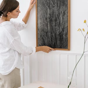 May include: A framed black and white art print depicting a close-up of tall, textured grasses. The artwork is in a light brown wooden frame. The image is being hung on a white wall.