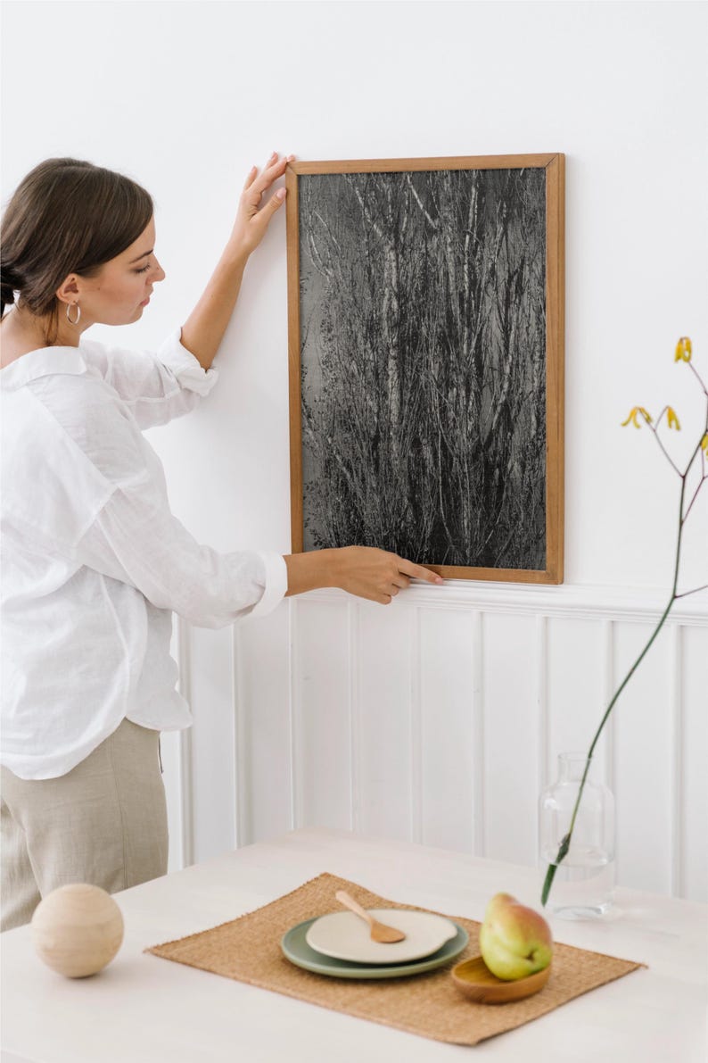 May include: A framed black and white art print depicting a close-up of tall, textured grasses. The artwork is in a light brown wooden frame. The image is being hung on a white wall.