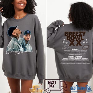 May include: A gray sweatshirt featuring a graphic of Chris Brown on the front. The back of the sweatshirt displays "Breezy Bowl XX" tour details, including European and North American tour dates. The sweatshirt is a unisex garment.