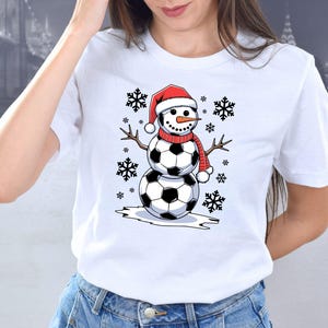 May include: White t-shirt with a snowman wearing a Santa hat and soccer balls for a body. The snowman is surrounded by snowflakes.