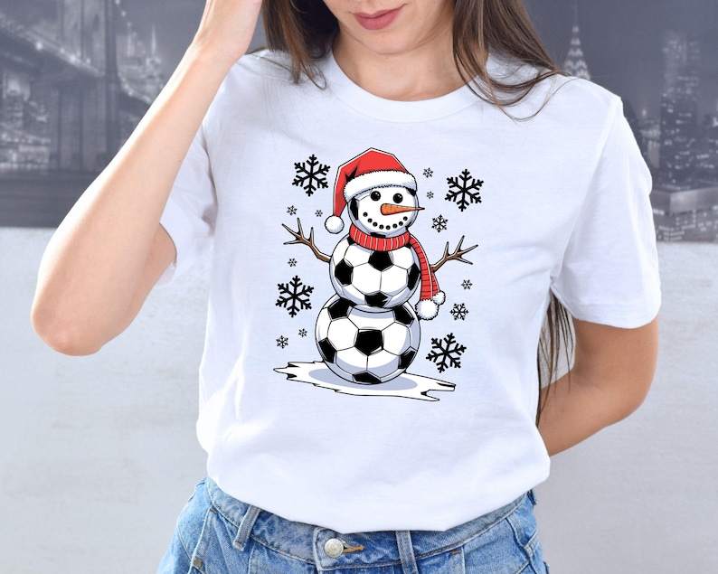 May include: White t-shirt with a snowman wearing a Santa hat and soccer balls for a body. The snowman is surrounded by snowflakes.