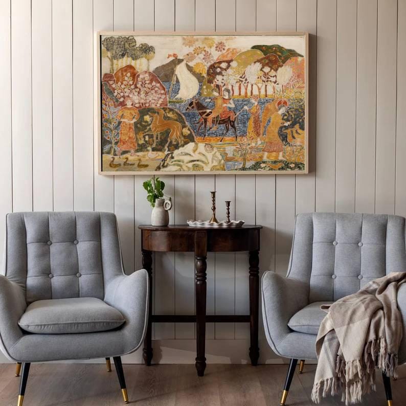 May include: A framed artwork with a colorful, abstract design hangs above a small, dark wooden table. Two gray armchairs with button tufting and gold-tipped legs flank the table. A neutral-toned throw blanket rests on one chair.