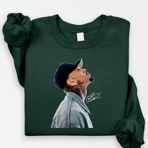 May include: Dark green sweatshirt featuring a portrait of a person wearing a denim jacket and a navy blue baseball cap, looking upwards. The portrait includes a signature in white. The sweatshirt has long sleeves.