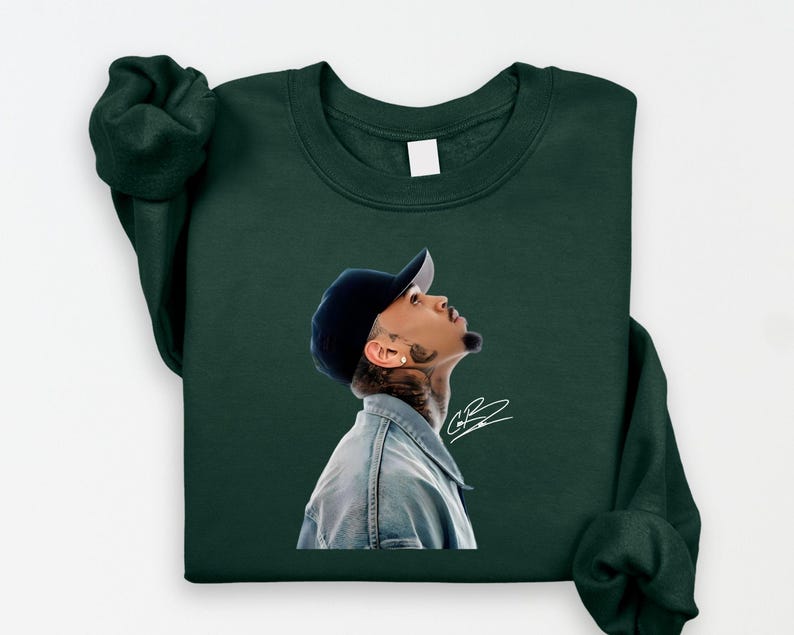 May include: Dark green sweatshirt featuring a portrait of a person wearing a denim jacket and a navy blue baseball cap, looking upwards. The portrait includes a signature in white. The sweatshirt has long sleeves.