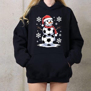 May include: Black hoodie with a snowman made of soccer balls wearing a Santa hat. The snowman is surrounded by snowflakes.