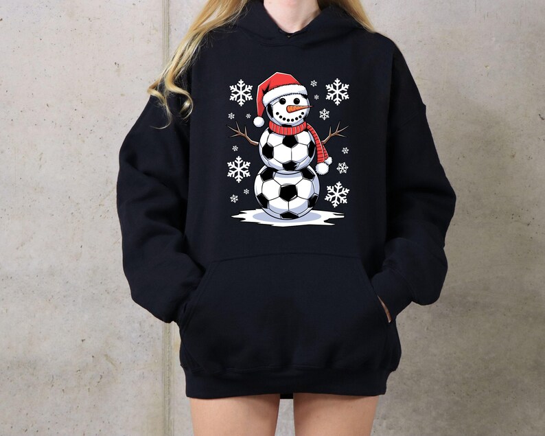 May include: Black hoodie with a snowman made of soccer balls wearing a Santa hat. The snowman is surrounded by snowflakes.