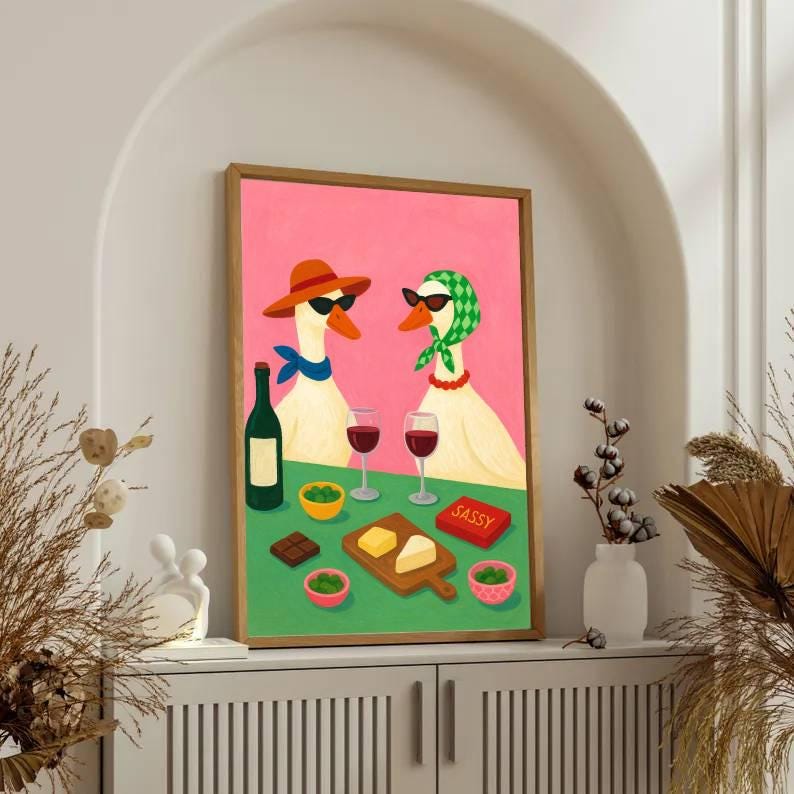 May include: A framed art print featuring two geese wearing sunglasses and accessories, enjoying wine and cheese. The artwork has a pink background and a green table with food and a wine bottle. The word "SASSY" is on a red block.