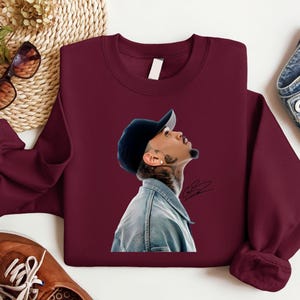 May include: Burgundy crewneck sweatshirt featuring a portrait of a person wearing a denim jacket and a black baseball cap. The portrait is on the front of the sweatshirt. Sunglasses and brown leather shoes are visible in the background.
